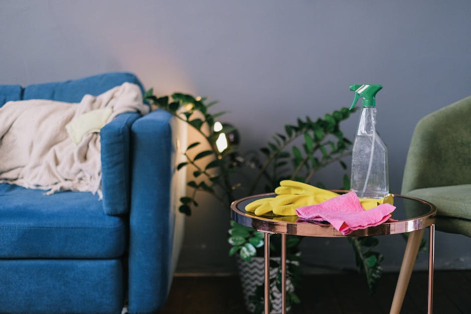 A person dressed in full white protective coveralls, including a hood and a face mask, is performing surface cleaning on a black fabric sofa in a well-lit living room. The individual is wearing blue g